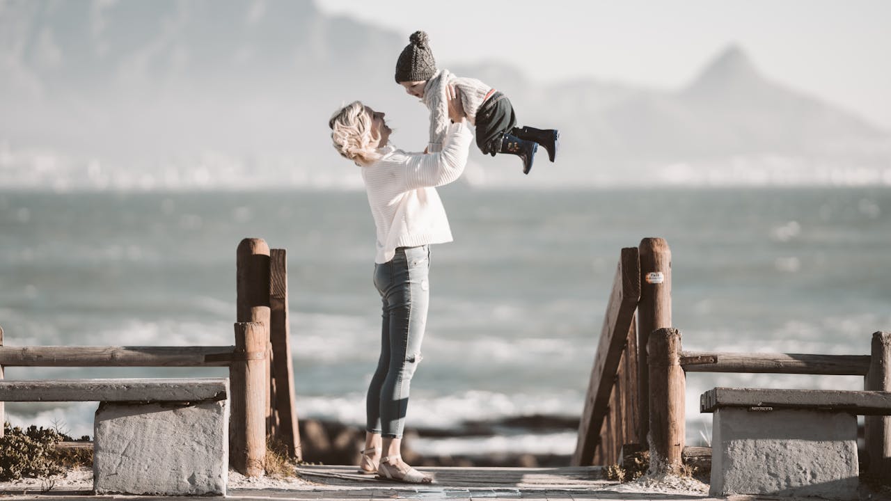 A joyful mother lifting her child near the beach on a sunny day.